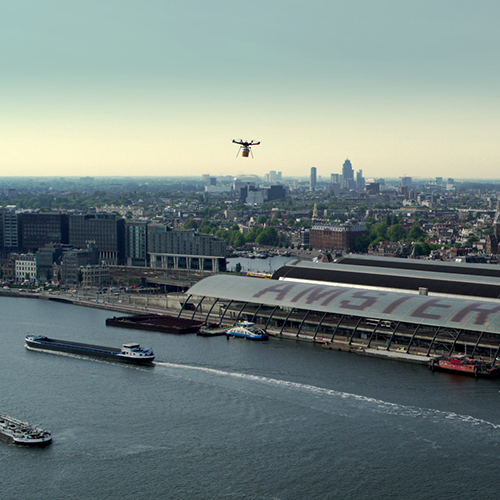 Thumbnail for the promo video Hilton Haring Party. An aerial shot overlooking Amsterdam Central Station. A drone is seen approaching the station.