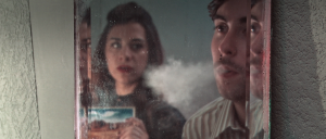 Still from the film Nowhere Place. The writer and the girl are seen in the reflection of a mirror. The writer blows smoke on the mirror and the girl is holding a postcard of a landscape.