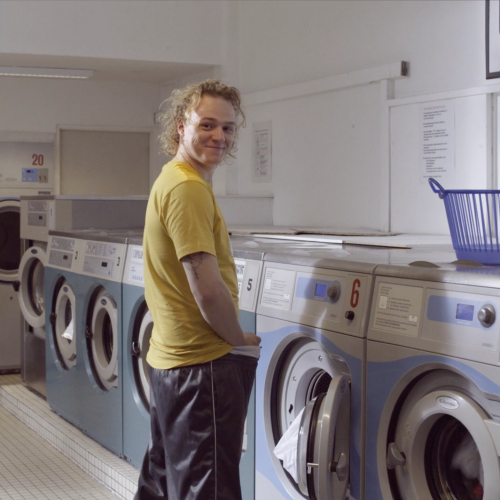 Still of the commercial Wasserette. A guy is standing in front of a washing machine in a laundrette. He's looking back and smiling.
