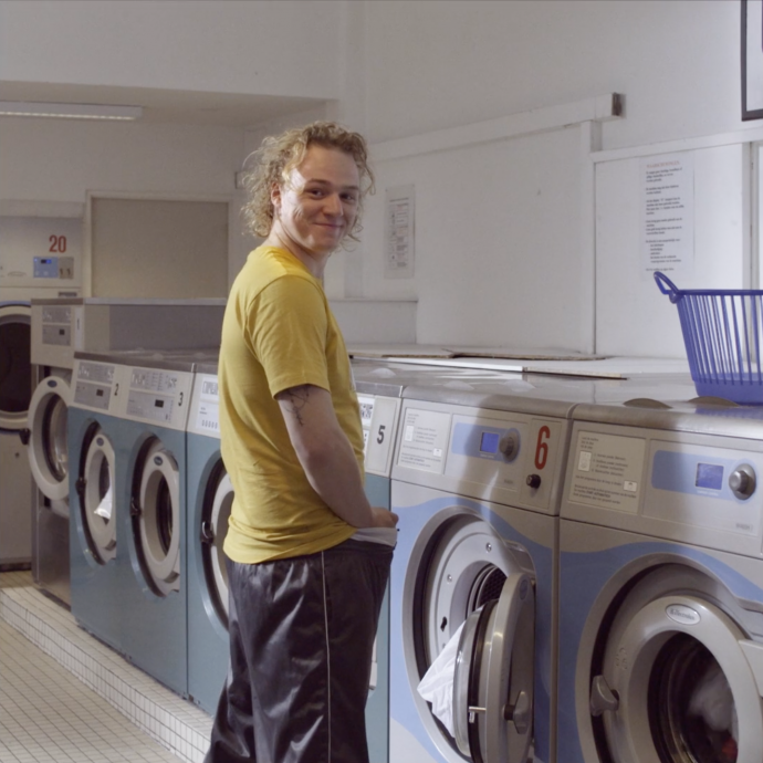 Still of the commercial Wasserette. A guy is standing in front of a washing machine in a laundrette. He's looking back and smiling.