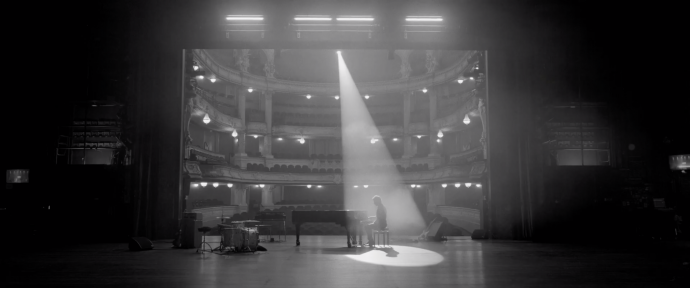 Still from the music video for Maarten Heijmans & Band - Sammy. Maarten is sitting behind a grand piano on the stage of the International Theater Amsterdam. There's a spotlight on him.