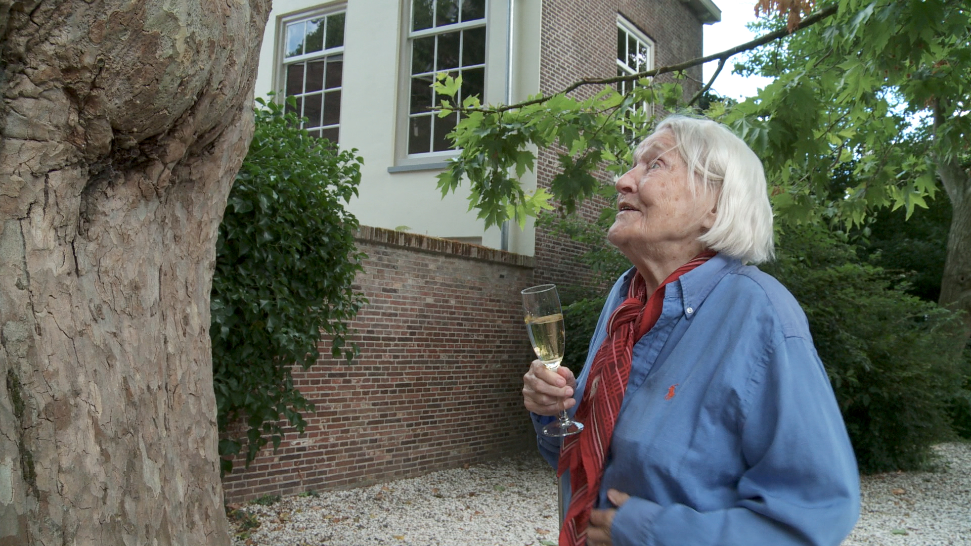 Still from the film Een klein beetje nog. Elisabeth is standing at an old tree with a glass of champagne in hand. She looks happy.