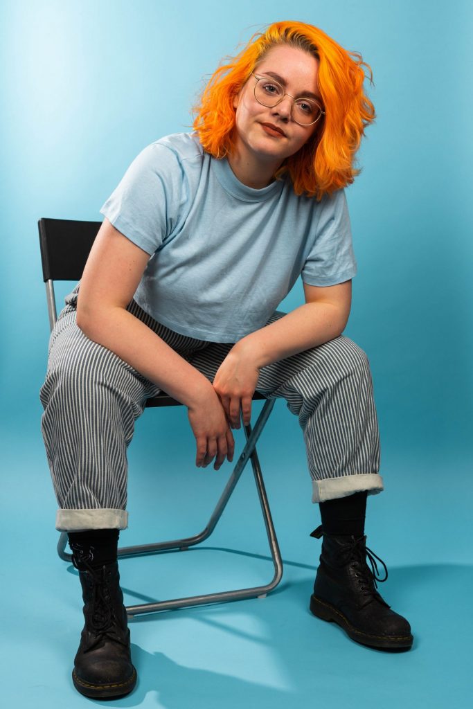 Picture of Eva sitting on a black fold-out chair in a light blue studio space. She's has bright orange hair, blue eyes and is wearing thin, gold-rimmed circular glasses. She's wearing a light blue cropped t-shirt, blue-and-white striped jeans and Dr Marten shoes. She's looking into the camera with a confident yet soft look.