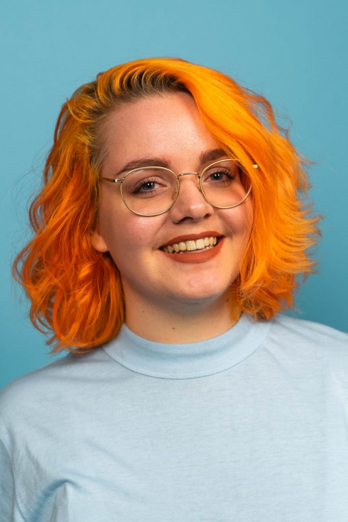 Headshot of Eva. She's pictured in front of a light blue backgroundand wearing a lighter blue shirt. She has shoulder length, slightly curly, bright orange dyed hair and blue eyes. She's wearing gold rimmed circular glasses.