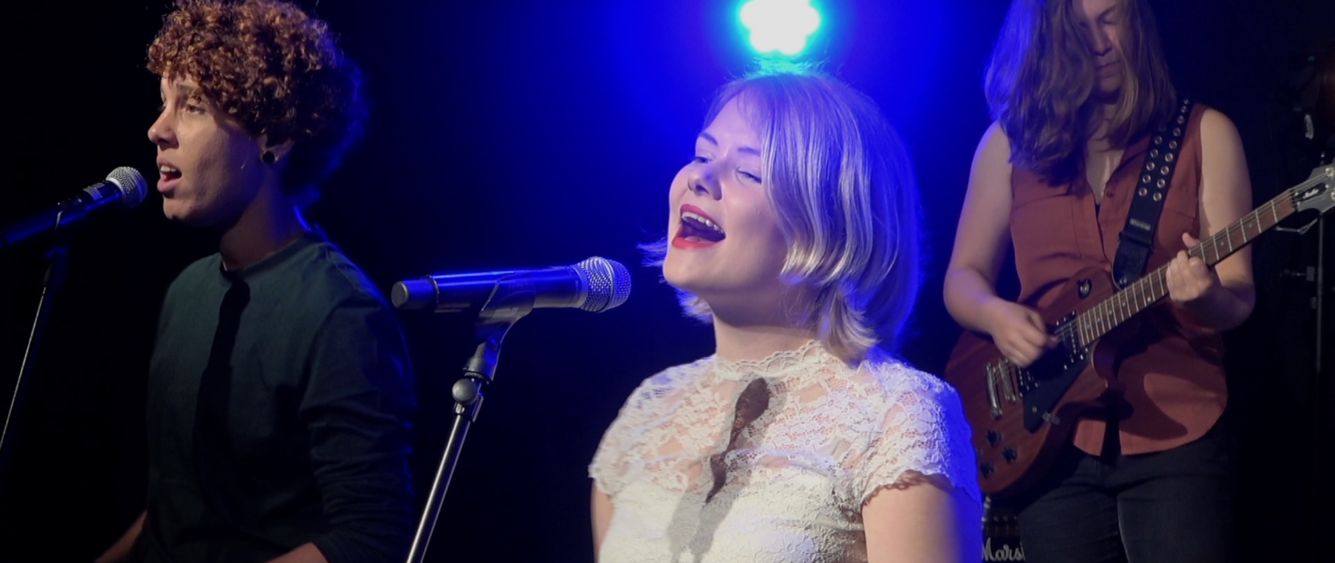 Still from Groei - Suzanne de Jong. Suzanne is standing in the middle of the frame in front of a microphone, singing. She has shoulder length blonde hair, wears a white lacy top and is wearing red lipstick. In the background is a back-up singer with short curly hair and a guitarist with long brown hair.