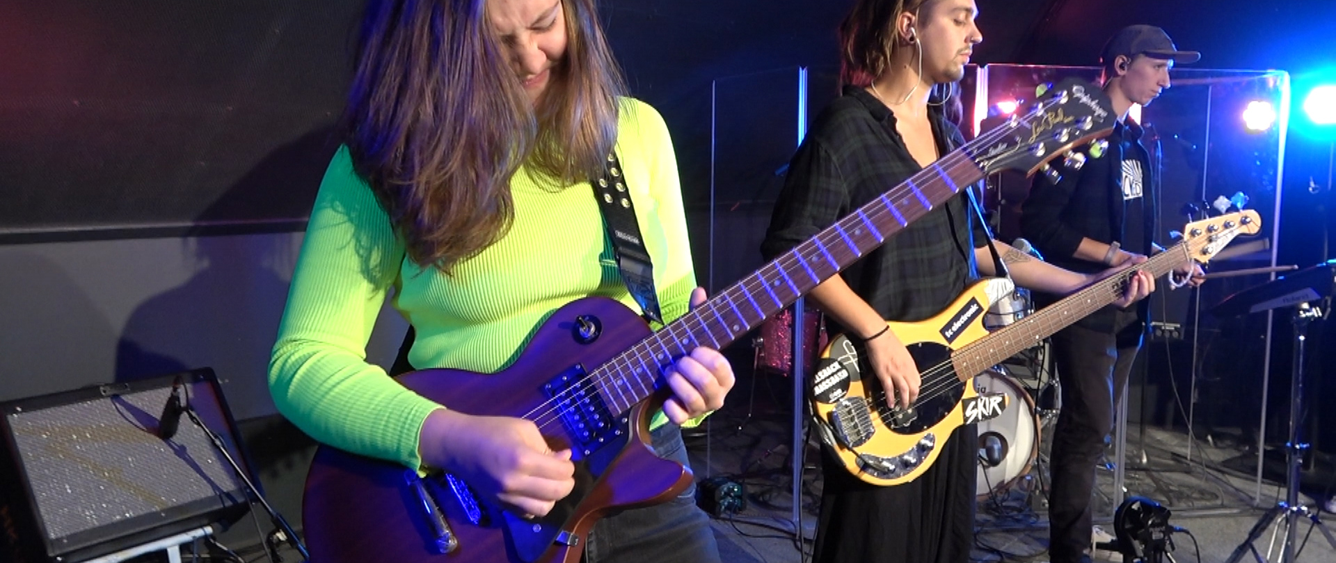 Still from Groei - Suzanne de Jong. A close-up shot of a guitarist. She is wearing a bright neon yellow/green top. Behind her is a bassist and a drummer.