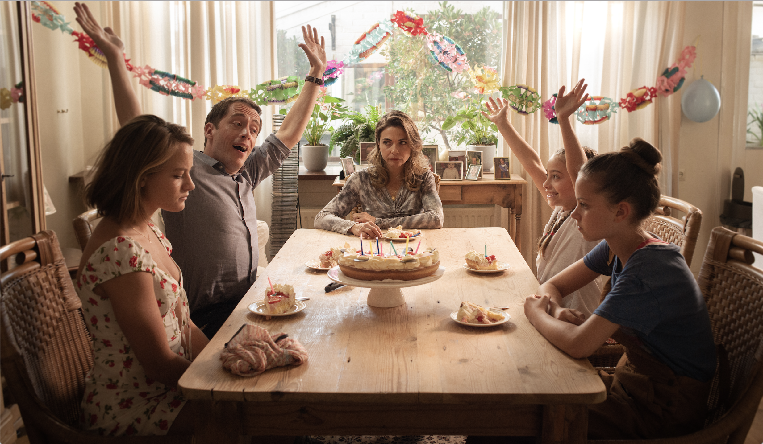 Still from Buiten is het Feest. The family is sitting at the dining table; it's someone's birthday. The mom in the middle, Willemke and Hannah are looking grumpy while Kees and Marta have their hands in the air.