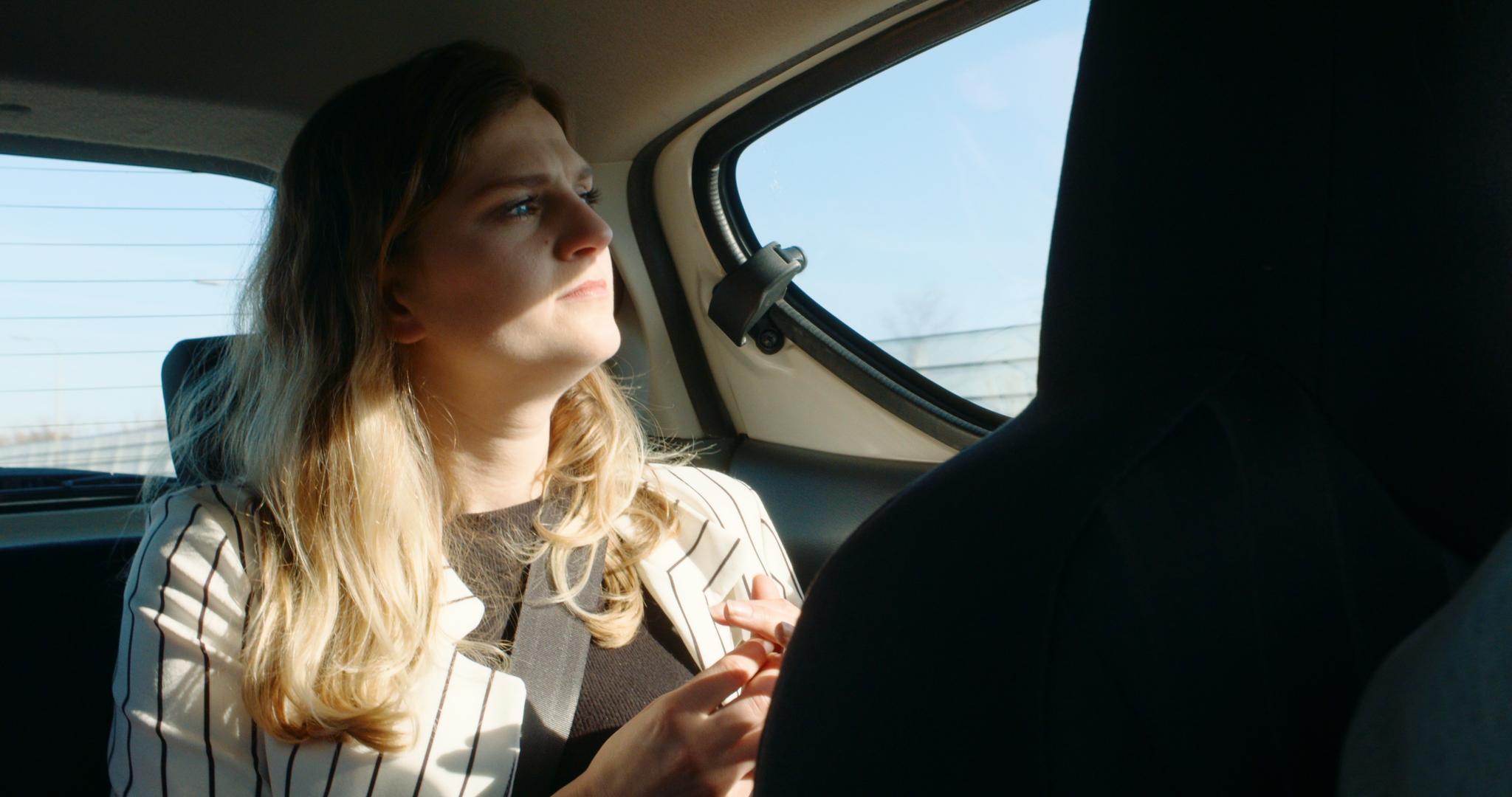 Still from Momentum. Carline is sitting in the back seat of a car and staring out of the window pensively. She's wearing a black top with a white pinstriped blazer. It's sunny.