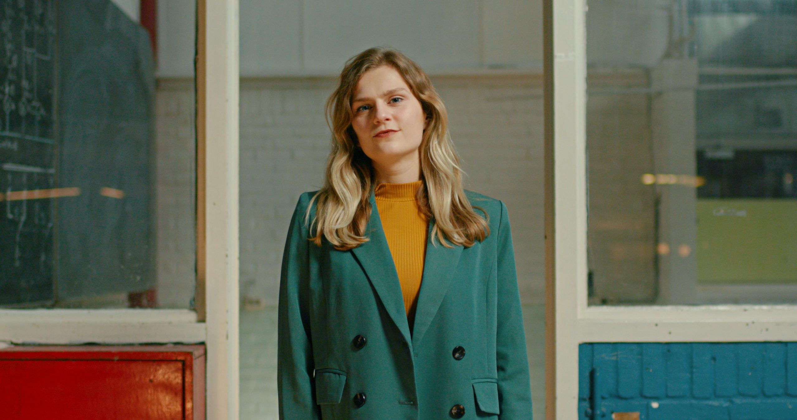 Still from Momentum. A medium of Carline standing in front of a doorway in what looks like an abandoned school. She's wearing a yellow top and a blue blazer, and she's staring powerfully into the camera.