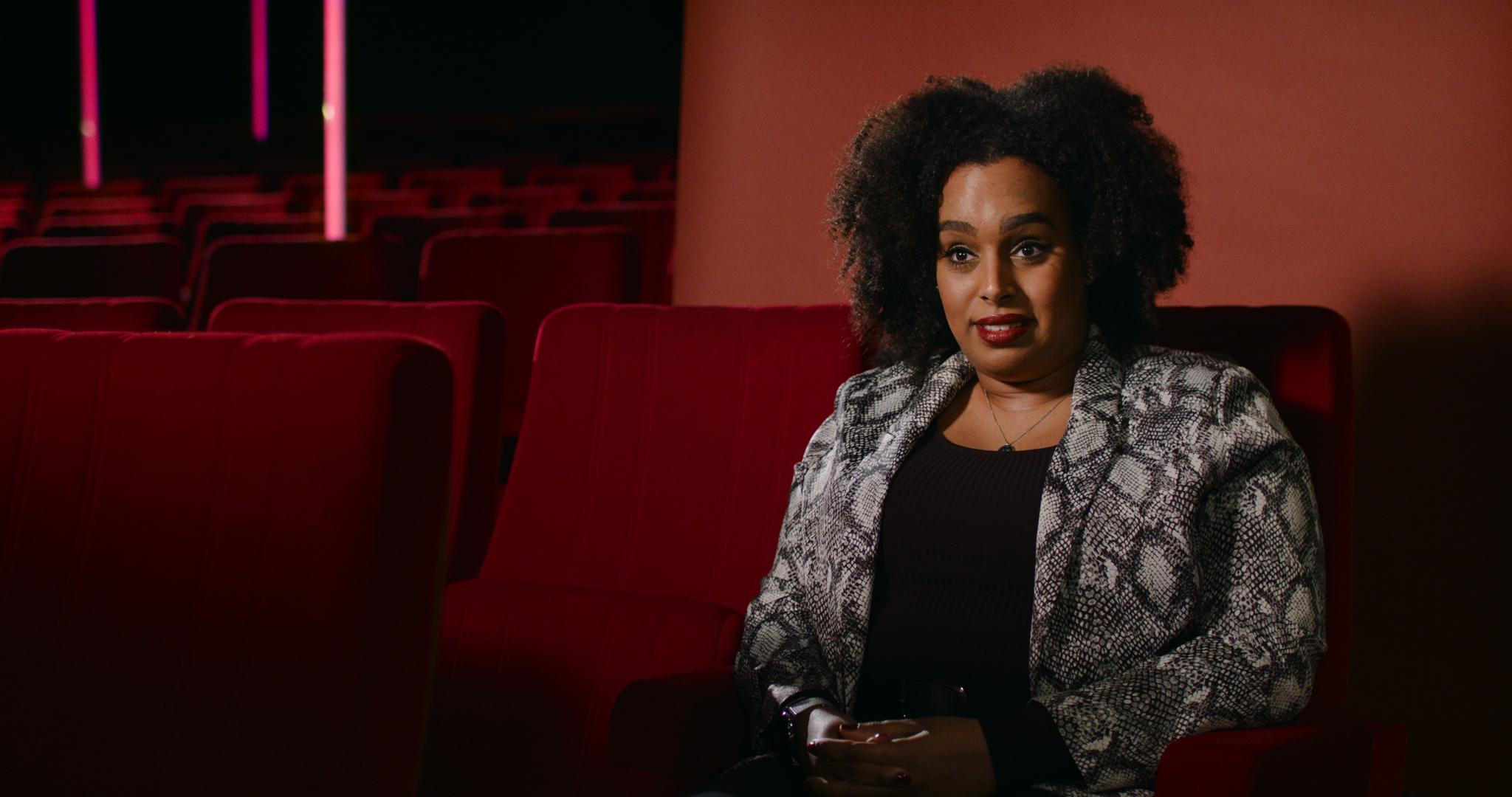 Still from Momentum. Mikal is sitting in a cinema chair with a red background set up; it's obviously in an interview setting.