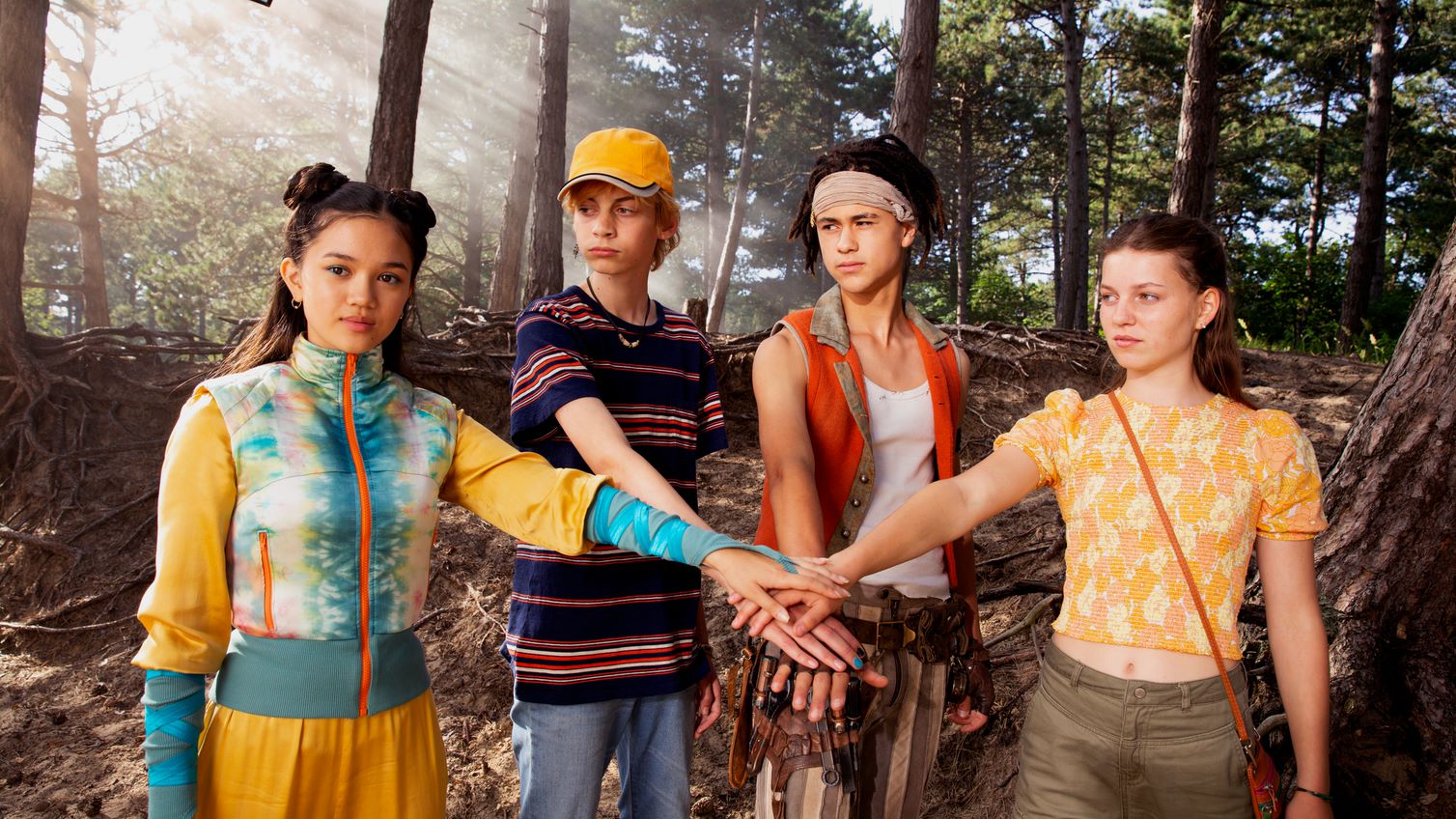 Still from De Piraten van Hiernaast 2. Yuka, Michiel, Billy and Elizabeth are standing in a forest with their hands outstretched, meeting in the middle, like they're going to do a cheer.