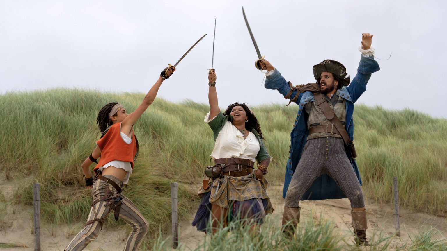 Still from De Piraten van Hiernaast 2. Billy, his mom Betsie and his dad Hector are standing on a dune, swords up in the air and cheering.. The swords are crossed in the middle.