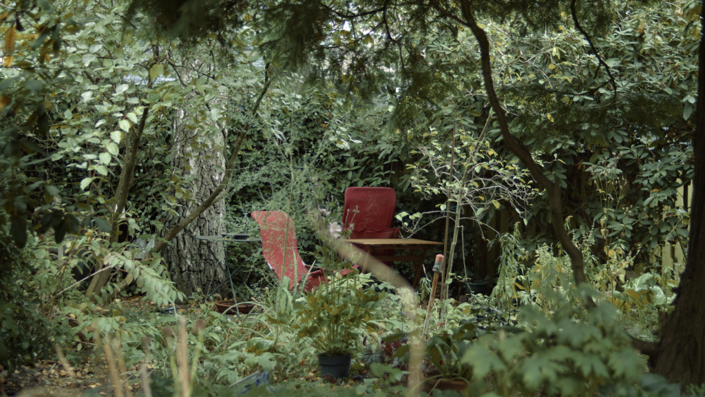 Still from the film 'Synchroon'. It's a wide shot of a seemingly overgrond garden with two empty red garden chairs in the centre. One of the chairs is folded shut against a garden table.