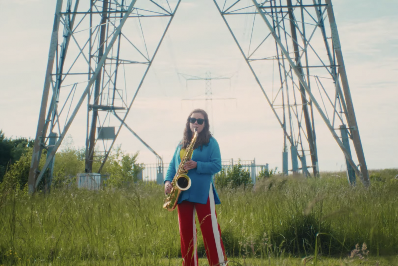 Still from 'Kom Van Dat Gas Af'. A young woman with long brown wavy hair is playing the saxophone in front at the foot of an electricity tower. She's wearing black sunglasses, a blue coat and red sporty type trousers.