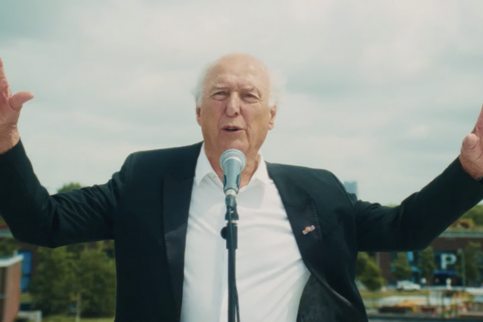 Still from 'Kom Van Dat Gas Af.' Peter Koelewijn, an old white man wearing a white blouse and a black suits jacket, is singing into a microphone in our direction. He has his arms wide open.