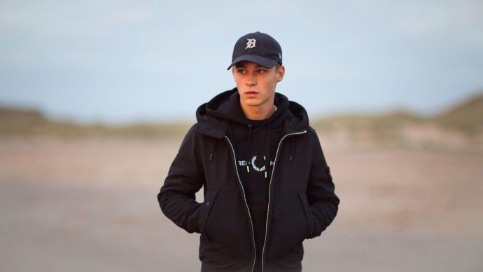 Still from the film 'Mascotte'. Jerry, a white teenage boy, is standing on a Dutch beach with the dunes behind him. He's wearing a black sweater, a black cap and a black thick winter coat. He's got his hands in his pockets and he looks stern.