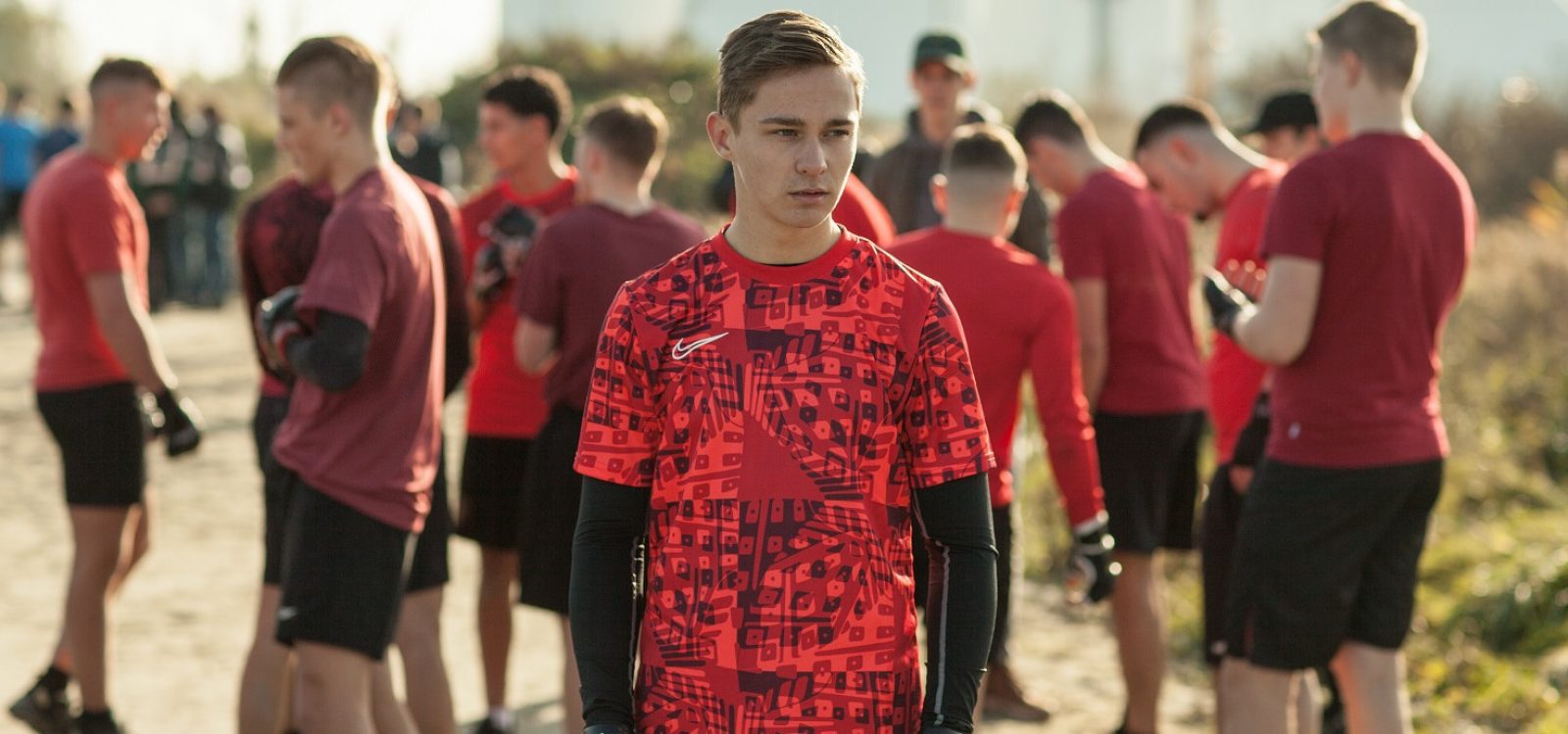 Still from the film 'Mascotte'. Jerry, a white teenage boy, is standing in the middle of the shot in red sports clothes. Behind him is a group of other teenaged boys, all wearing sports outfits with reds shirts.