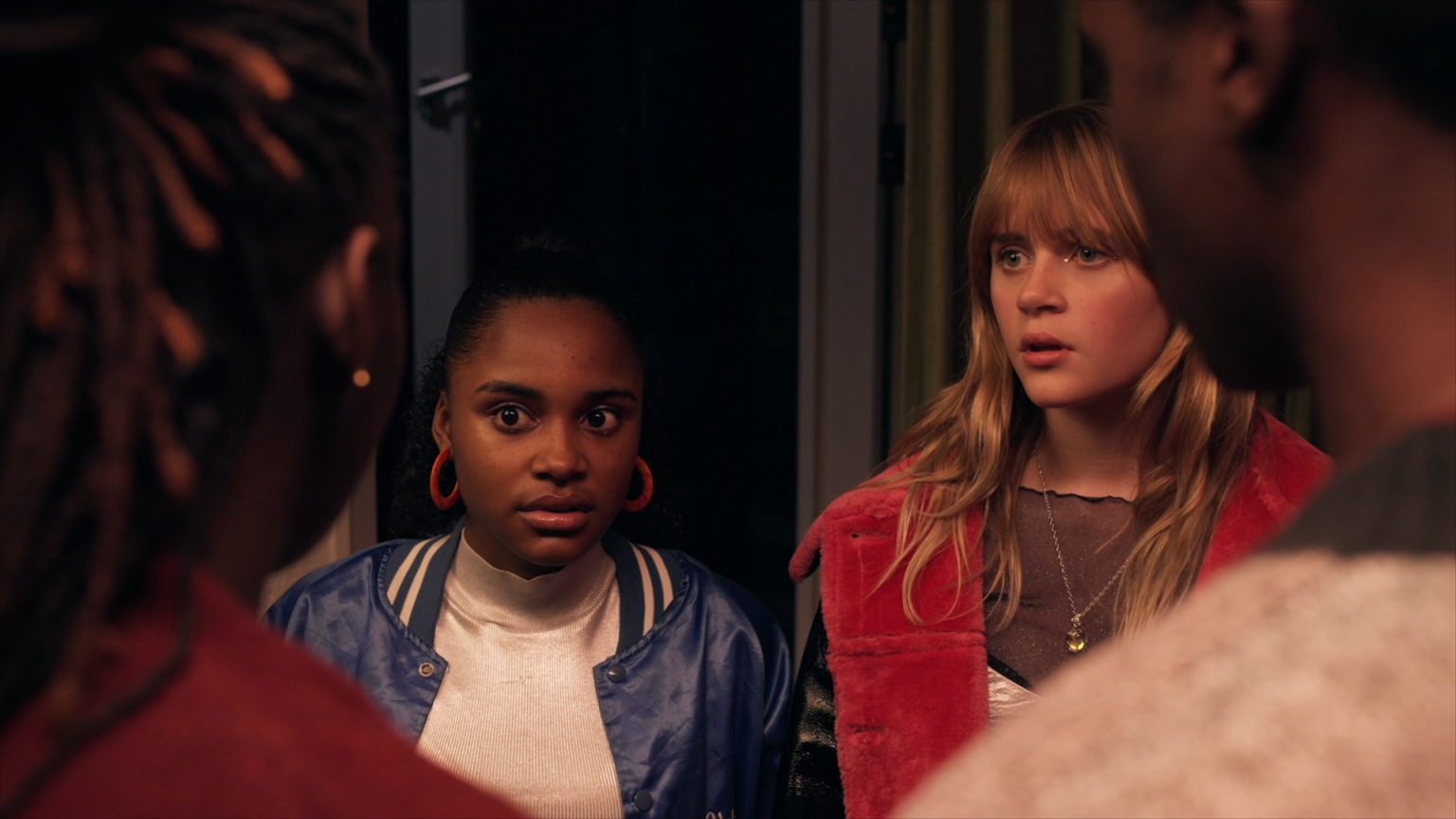 Still from 'Gedrogeerde Ouders?', episode 6 of Broodje Aap. It is an over the shoulder shot of two teenaged girls, one Black and one white, in hip 'going out' clothing. They are looking shocked at the two people we are looking over the shoulder from; two Black adults.