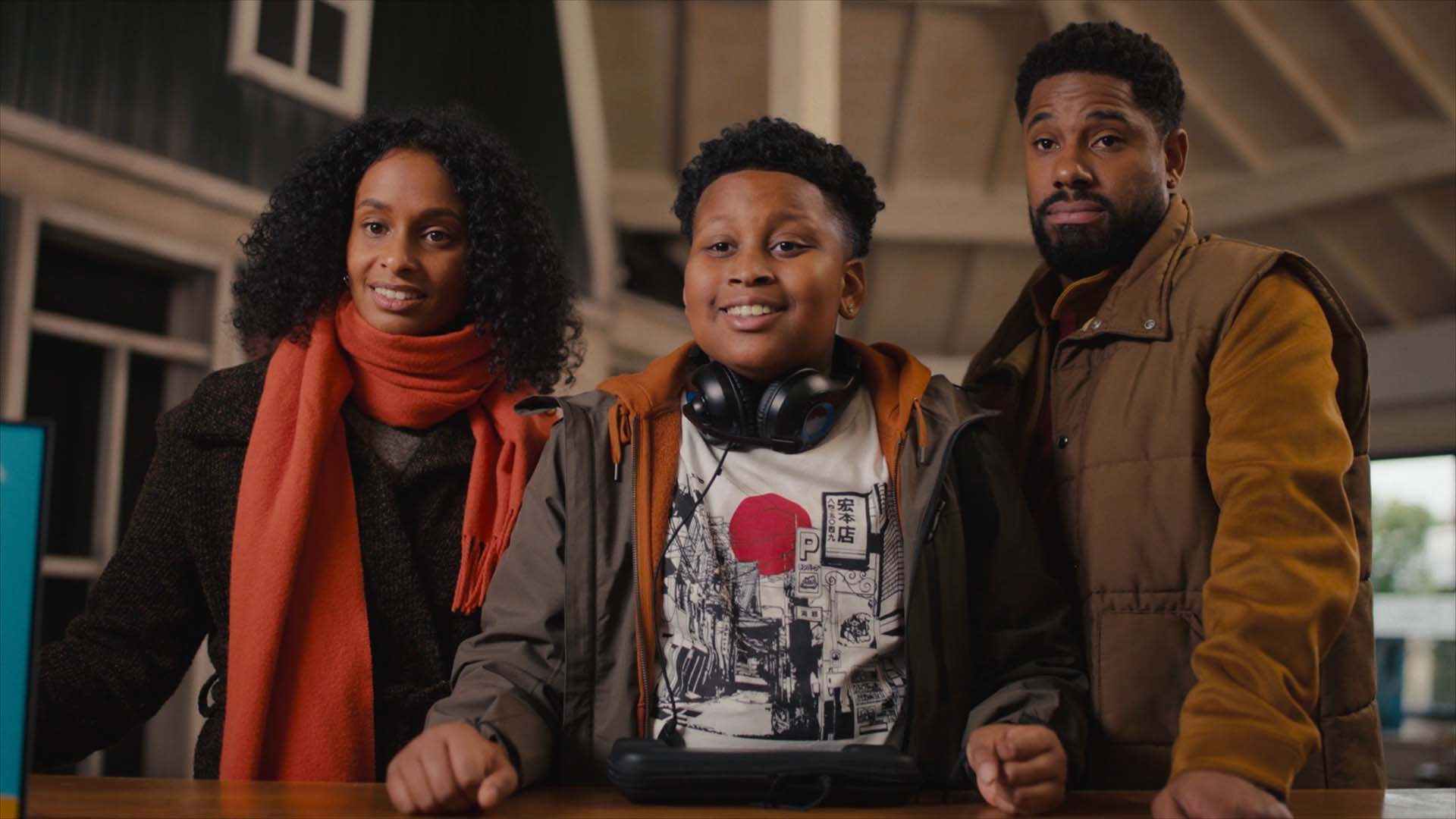 Still from 'Game Over', episode 5 of Broodje Aap. It's a medium shot of a Black family standing at a desk in a holiday park reception. From left to right: the mother, with a big head of curls and a red scarf; the teenaged son with a graphic t-shirt, jacket and headphones around his neck; the father, with a yellow-brownish jacket and body warmer. The mother and son are smiling, the father has his eyebrows raised in a questioning manner.