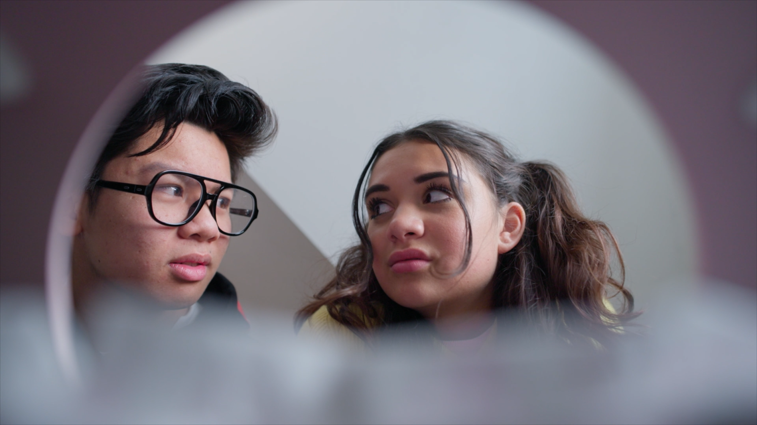 Still from 'Bizarre Date', episode 14 of Broodje Aap. It's a POV from within a toilet bowl. The foreground has some blurry toiletpapier. Hanging above the toiletbowl, looking at each other, are two teenagers (one asian boy with dark hair and thick rimmed sunglasses, and one white girl with brown hair in two pigtails).