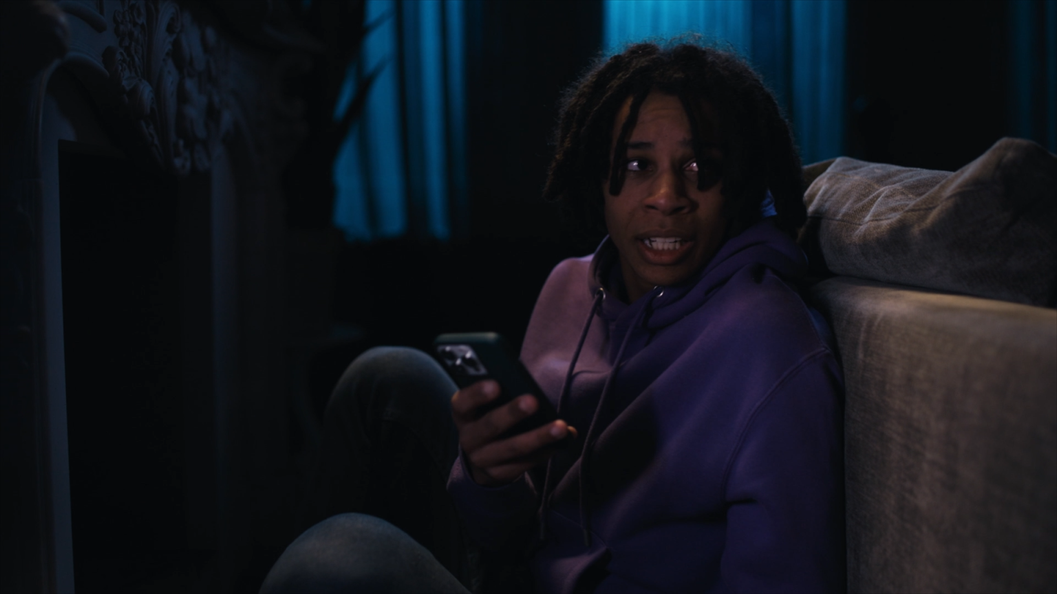 Still from 'Telefoonstalker', episode 13 of Broodje Aap. It's a medium shot of a Black teenaged guy, with braids and a purple hoodie, sitting behind a couch. He's holding his phone and looking anxiously over his shoulder. The room is dark.