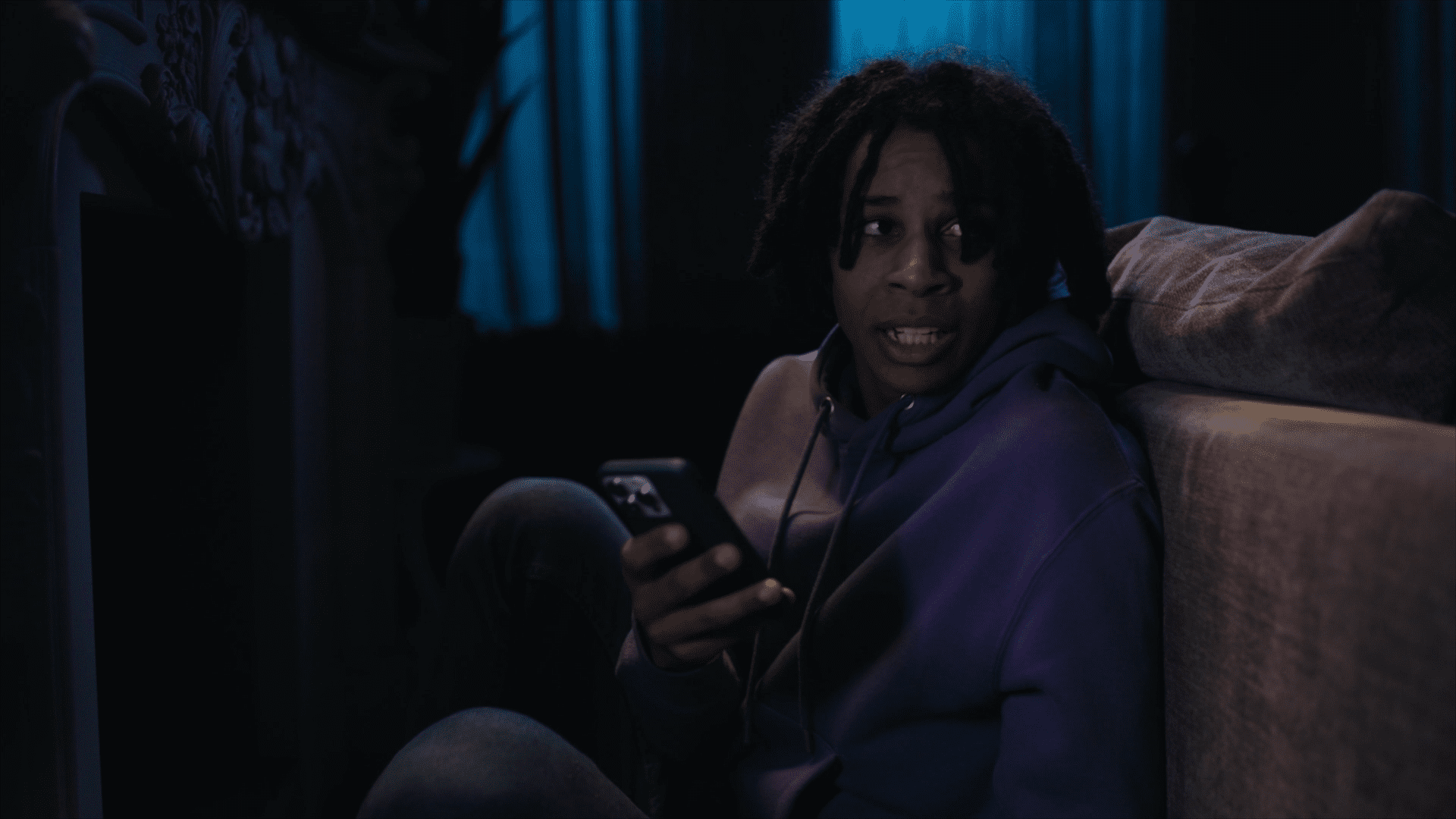 Still from 'Telefoonstalker', episode 13 of Broodje Aap. It's a medium shot of a Black teenaged guy, with braids and a purple hoodie, sitting behind a couch. He's holding his phone and looking anxiously over his shoulder. The room is dark.