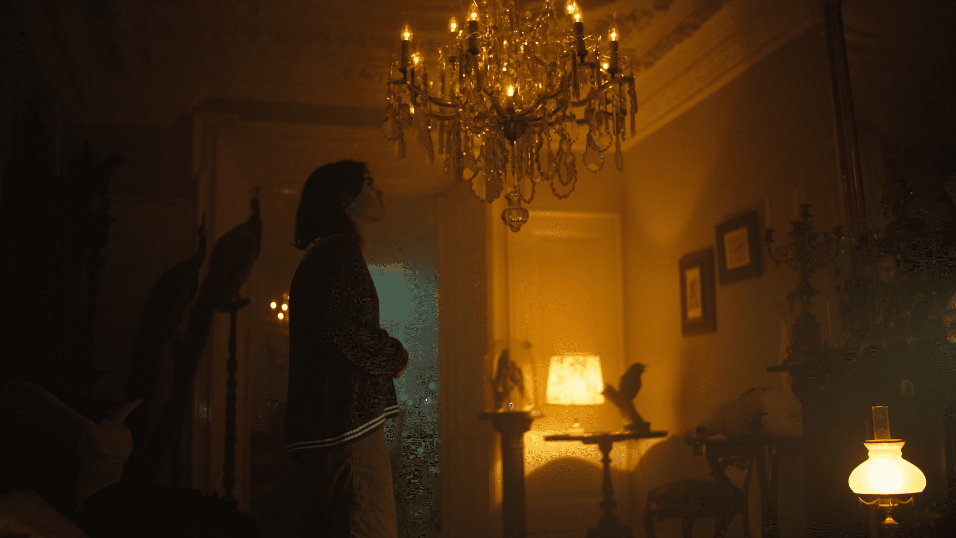 Still from 'Oppassen...', episode 8 of Broodje Aap. It's a wide shot of a teenaged girl standing in the middle of the living room of an old house. The lighting is moody and dark, and the girl is looking up at a big chandelier in the middle of the room. The vibe is creepy.