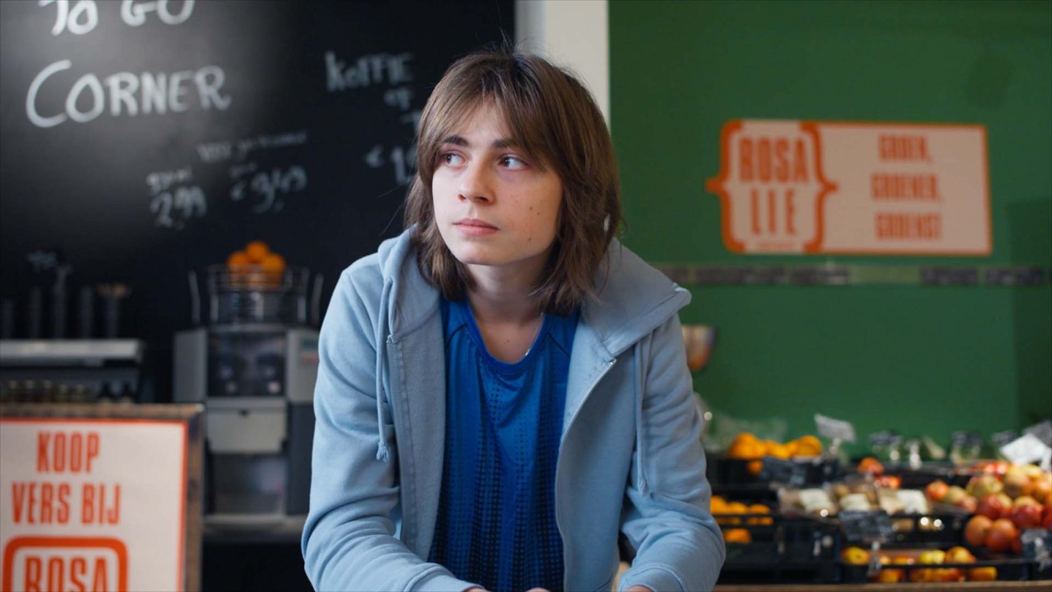 Still from 'Terror Zusje', episode 12 of Broodje Aap. It's a medium shot of a teenaged boy leaning on something off-screen in the middle of a grocery store. He has shoulder length light brown hair, a blue t-shirt and light blue hoodie and is looking bored, to the left.