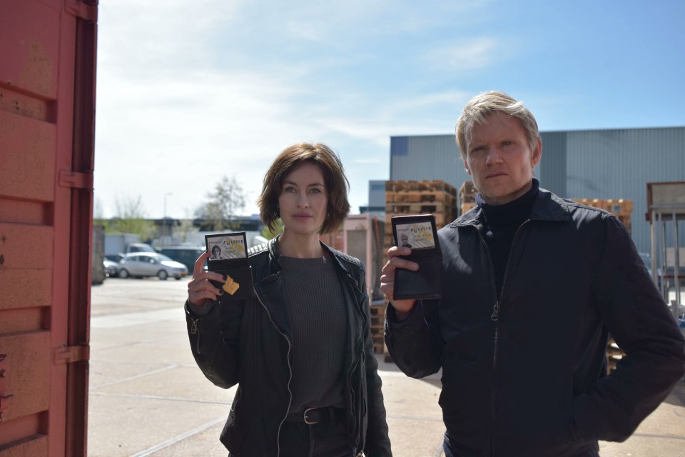 Still from Van Der Valk S3E1 'Freedom in Amsterdam'. Lucienne and van der Valk are standing in what looks like an industry-type environment with big containers around them. They are flashing their police badges to someone off screen.