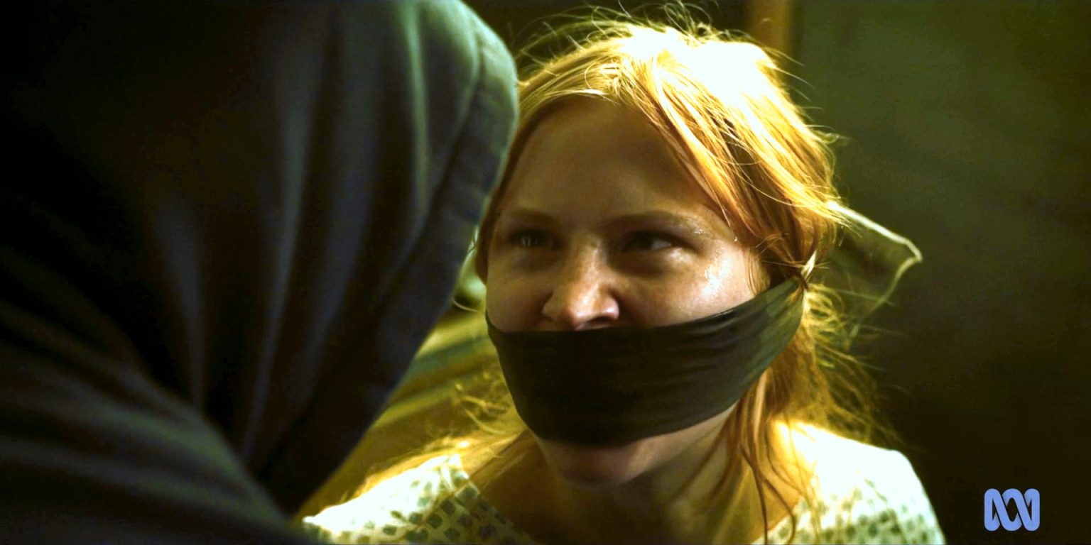 Still from Van Der Valk S3E1 'Freedom in Amsterdam'. A close-up shot of Marian Segers, a young ginger haired girl, who has a piece of cloth bound over her mouth, looks angrily at a hooded character, who we see over-shoulder.