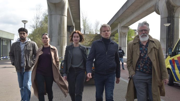 Still from Van Der Valk S3E1 'Freedom in Amsterdam'. The new crew walks underneath some Southeast Amsterdam-style metro infrastructure towards the camera in a medium wide shot. The characters are, frrom left to right: Eddie Suleman, Citra Li, Lucienne Hassell, Piet van der Valk and Hendrik Davie.