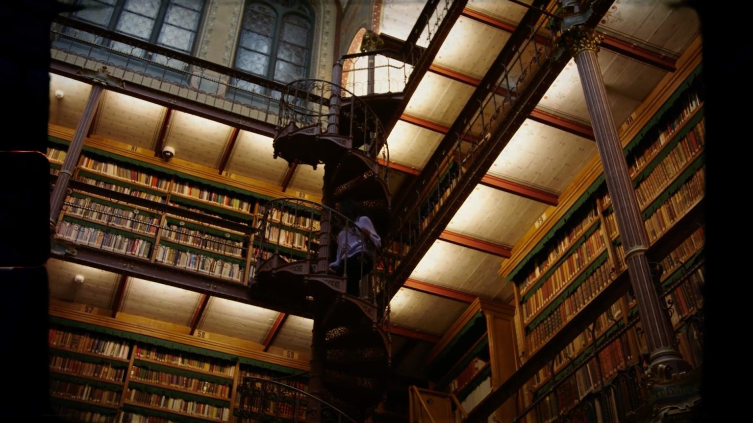 Still from Vaandeldrager - Rijksmuseum. A wide shot pointed up at a winding staircase in the middle of a huge library. A woman with a pastel lilac coloured sweater is walking up the stairs.