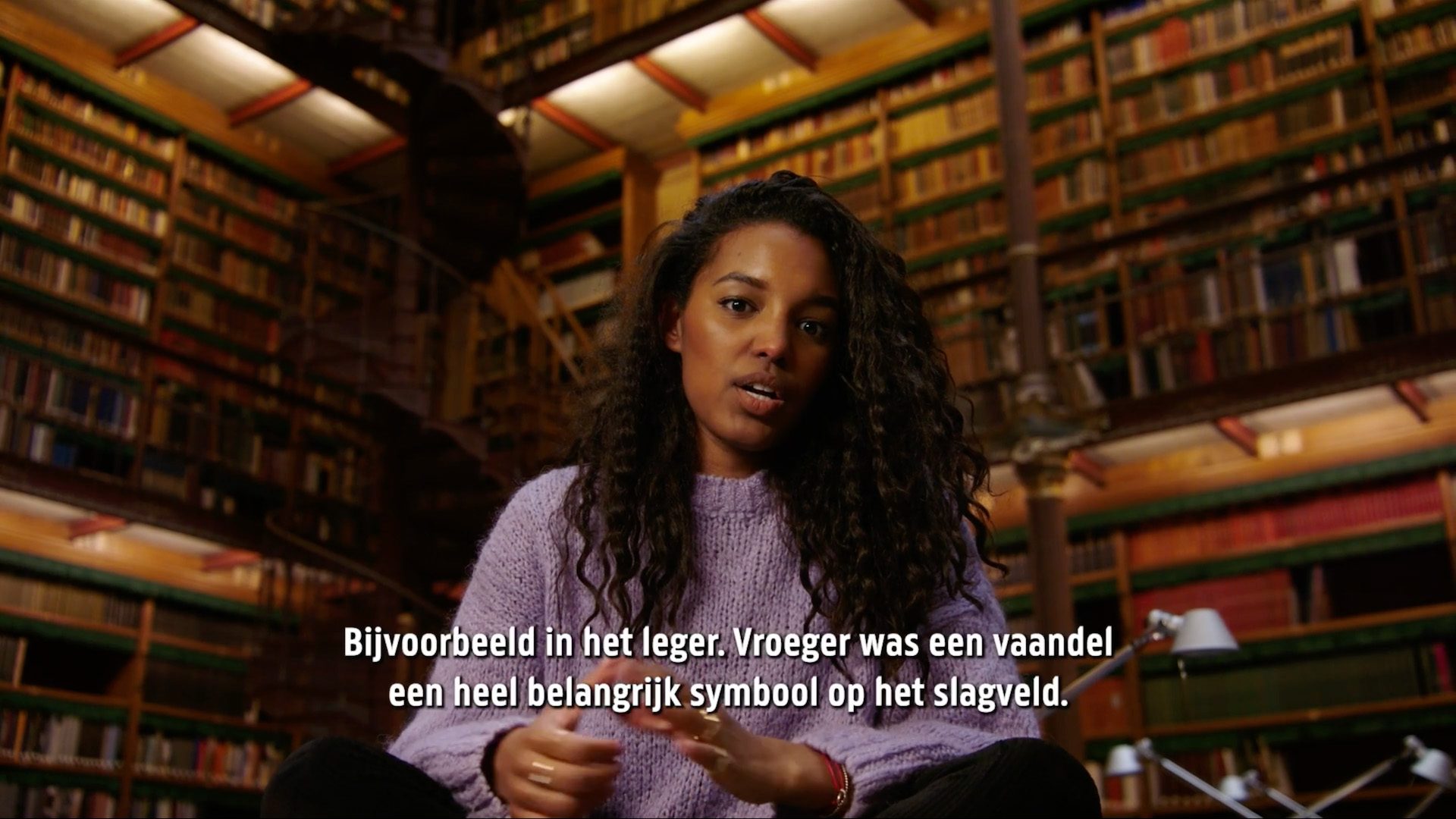 Still from Vaandeldrager - Rijksmuseum. A black woman with long, dark, curly hair, wearing a lilac sweater. She's sitting crosslegged (on a table off screen) in a dark but warm library. She is looking directly at the camera, and the image displays Dutch subtitles saying: "Bijvoorbeeld in het leger. Vroeger was een vaandel een heel belangrijk symbool op het slagveld.".