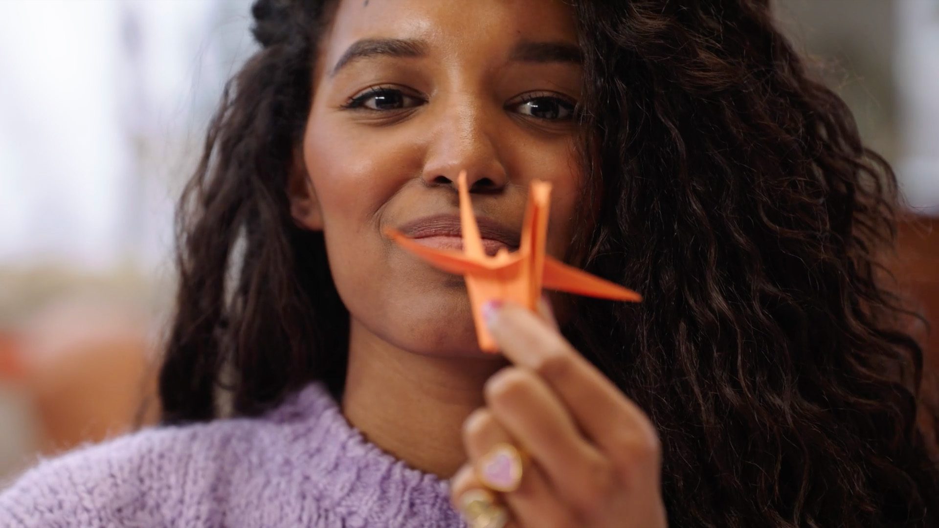 Still from Vaandeldrager - Rijksmuseum. A close-up of a black woman with long, dark, curly hair, wearing a lilac sweather. She is holding an orange paper crane in her fingers in front of her, her face in a pleased smile.