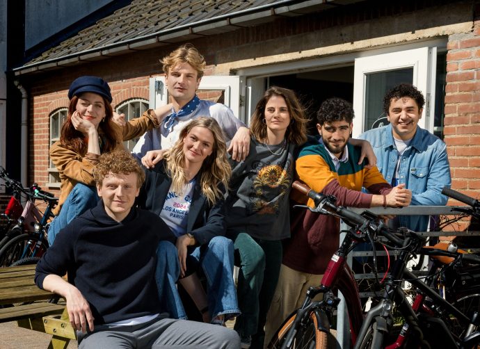 A group picture of the friend group from season 5 of Dertigers (Thirty-Somethings). They are posed outside, and are all looking at the camera. On the top row, from left to right: Ada, a white woman with red hair, wears a pageboy cap and is leaning her hand on Jonas, chin on her hand and an arm around Ruben; Ruben, a white man with blonde hair in a middle part, a white t-shirt and a blue handkerchief tied around his neck, leans his left arm on Suus; Suus, a white woman with brown hair, small silver hooped earrings and a colourful beaded necklace, has her arm around Reza; Reza, a man of Middle-eastern descent with curly dark brown hair and a beard, leans his arms on a fence-type structure; Sam, a man of Middle-eastern descent with shorter, curly dark hair, wearing a denim blouse. On the bottom row, from left to right: Jonas, a white man with curly, dark blonde hair and a black hoodie, is sitting on a bench of a picknick table; Laura, a white woman with blonde hair, jeans, t-shirt and a black blazer, is sitting on the table of the picknick table, with her arm resting on her knee.