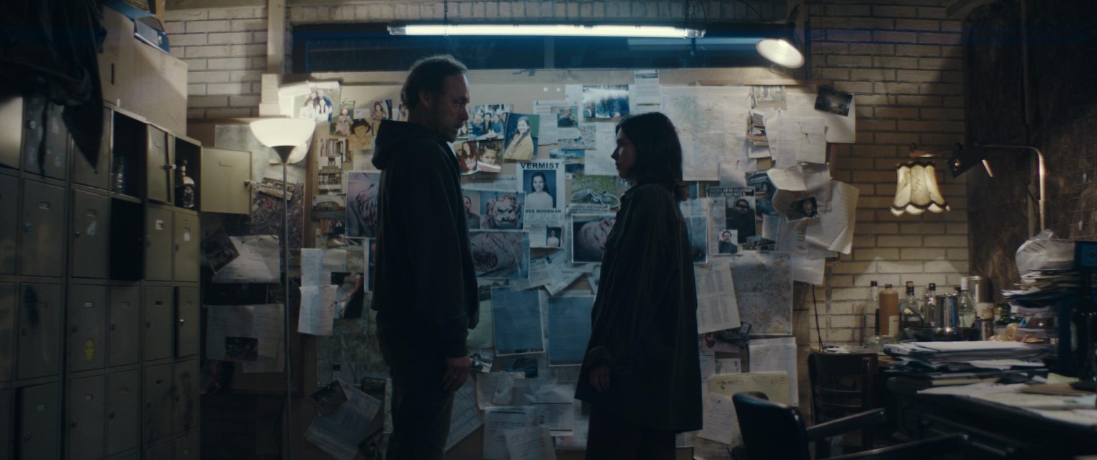 Still from Sphinx. Minke and her dad are standing in front of a huge wall of newspaper clippings, photos, posters, etc. In the centre is the 'Missing' poster of Minke. Minke and her dad are seen from en profil, with their faces to each other.