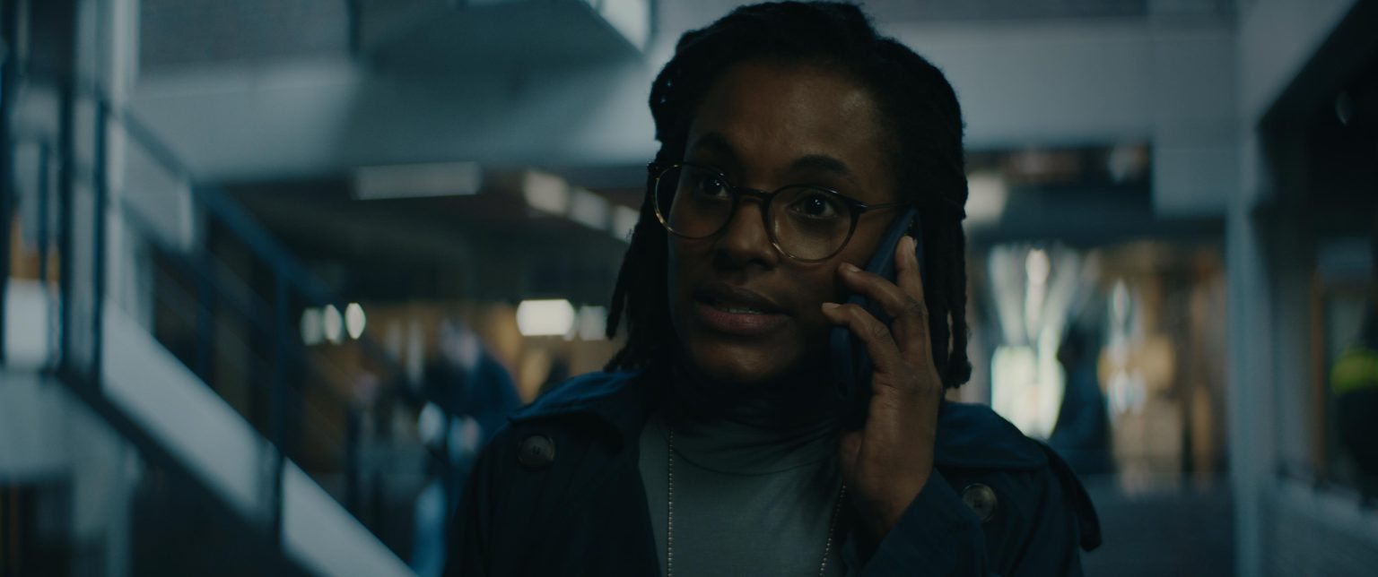 Still from Sphinx. A close-up still of Asha, a black woman with short braided hair and big rounded glasses. She is on the phone and her surroundings look like some kind of office/police station.