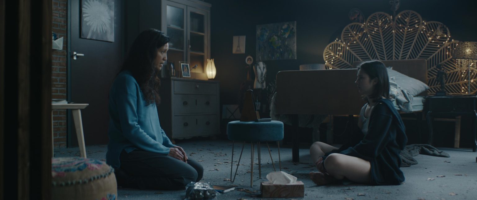 Still from Sphinx. This is a wide shot of Minke's bedroom. Minke is sitting cross legged on the floor on the right side of the frame, facing the left side. Across from her is an Asian woman with long black hair sitting on her knees. In between them is a stool with a metronome on it.