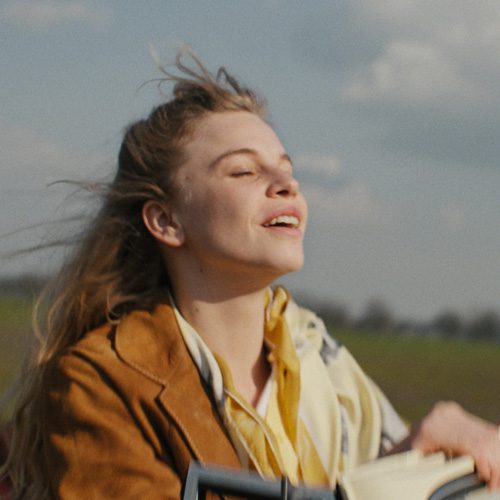 Still from Tegendraads. Johanna, a white Dutch woman with blonde hair, is standing up in 1960s open-top car. The wind is blowing through her hair and she has her eyes closed in a sort of wistful, content, happy relaxation.