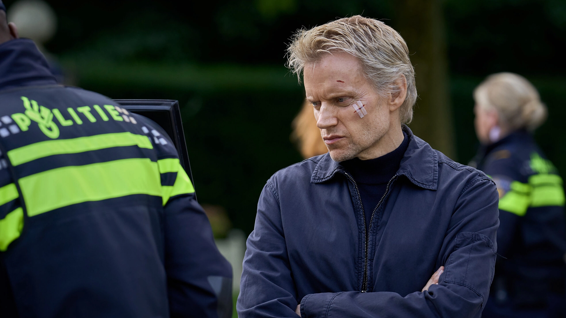 Still from Van der Valk S4 E1 'Safe in Amsterdam'. It's a medium shot on Piet van der Valk. He's looking to the left side, brows furrowed and arms crossed. On his cheek is a cut with a bandage on it. Visible on the fore- and background are the backs of two Dutch police officers.