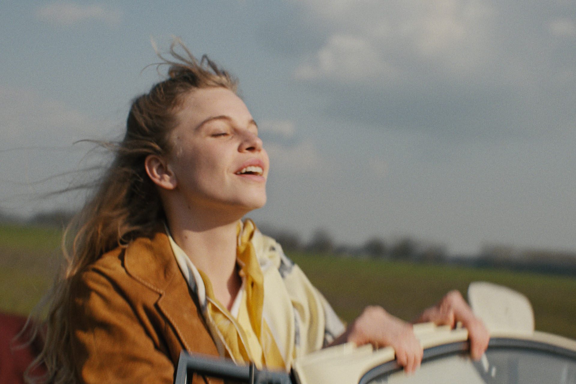 Still from Tegendraads. Johanna, a white Dutch woman with blonde hair, is standing up in 1960s open-top car. The wind is blowing through her hair and she has her eyes closed in a sort of wistful, content, happy relaxation.