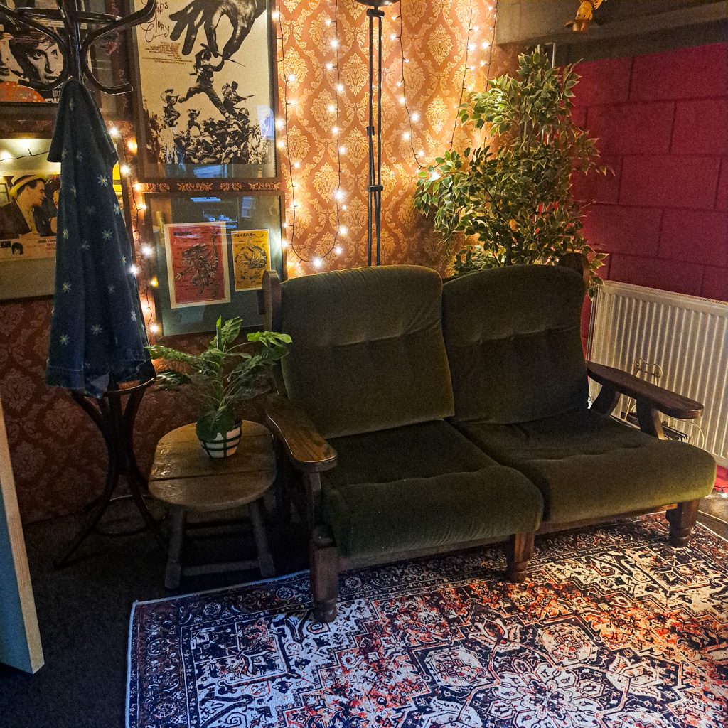 Picture from Eva's editing studio. The whole room feels vintage/thriftshop vibes. Pictures on the wall, a vintage green coach, fake plants, a 'persian' rug, an old brown café style coat rack. On the back wall are fairy lights.