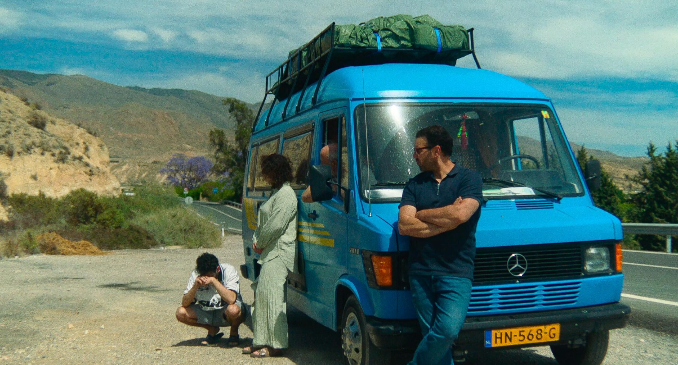 Still from Porte Bagage. It's a wide shot of a blue van parked to the side of the road, somewhere in the south of Europe. Leaning against the hood is Farid, wearing a short sleeved dark navy t-shirt and jeans. Leaning to the side of the van is Noor, with her hands clamped in front of her. Her face isn't visible. Crouching on the ground with his head in his hands is Hamza, a young Dutch-Moroccan man with a white t-shirt and jean shorts.