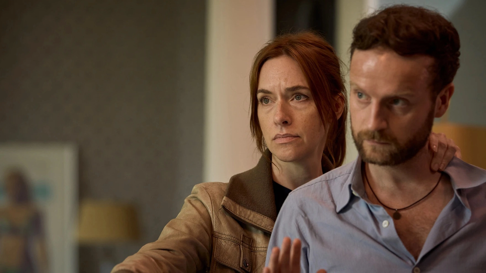 Still from Van Der Valk S4E1 Safe in Amsterdam. We see detective Van der Valk's former flame Kalie, a white woman with reddish brown hair behind a white man with short brown hair and a short beard.