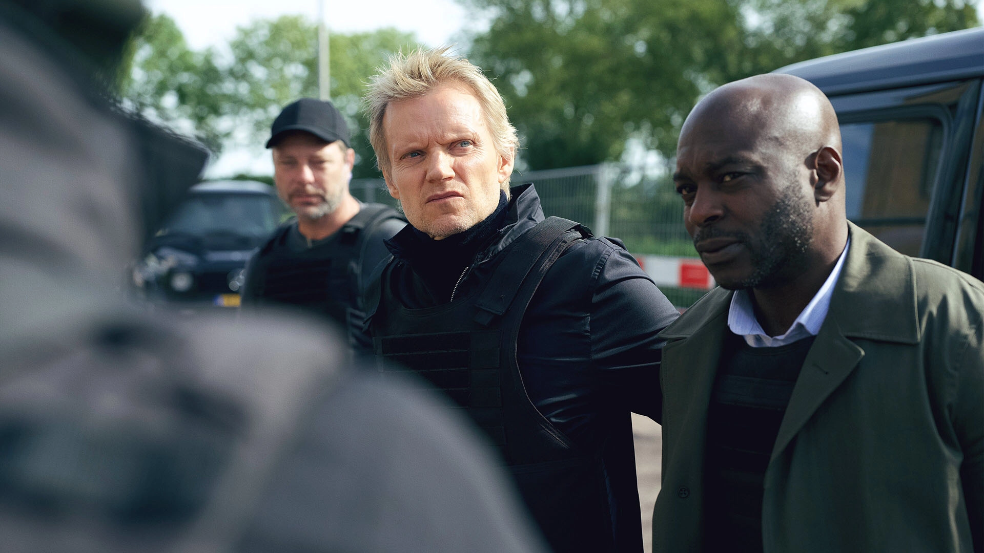 Still from Van der Valk S4E1 Safe in Amsterdam. Over-the-shoulder medium shot on Van der Valk, slightly turned away from the camera. We're outside, there's black vans behind him. He's looking pensively to the upper right corner. Flanked on either side are two more men; a white middle-aged man on his right side (our left) wearing a black cap and a bulletproof vest, a bald, black man on his left (our right) wearing a collared shirt with a vest and a thin coat over it.