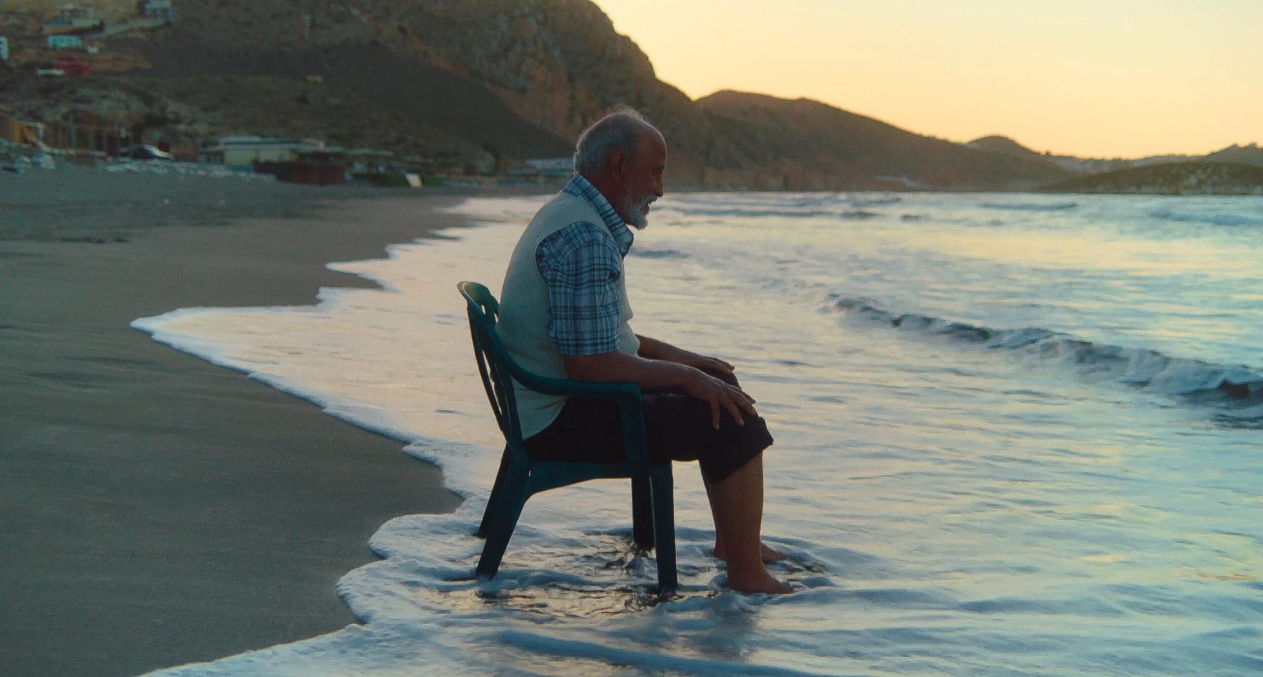 Still from Porte Bagage. A medium-wide shot of Musa Idrissi sitting on a plastic chair on the beach. His chair (and feet) are in the water, and the sun is setting.