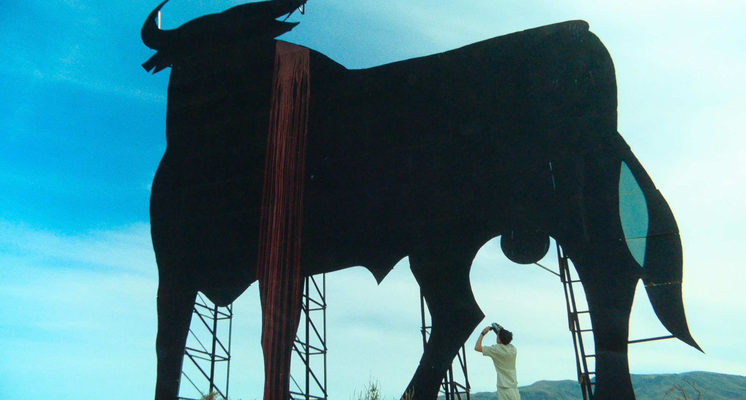 Still from Porte Bagage. A wide shot of a huge, black billboard in the shape of a bull against a blue sky with some clouds. In the bottom right you can see Rayan, a Dutch-Moroccan teenager. He wears a white t-shirt and is filming the billboard on a 1990s camcorder.