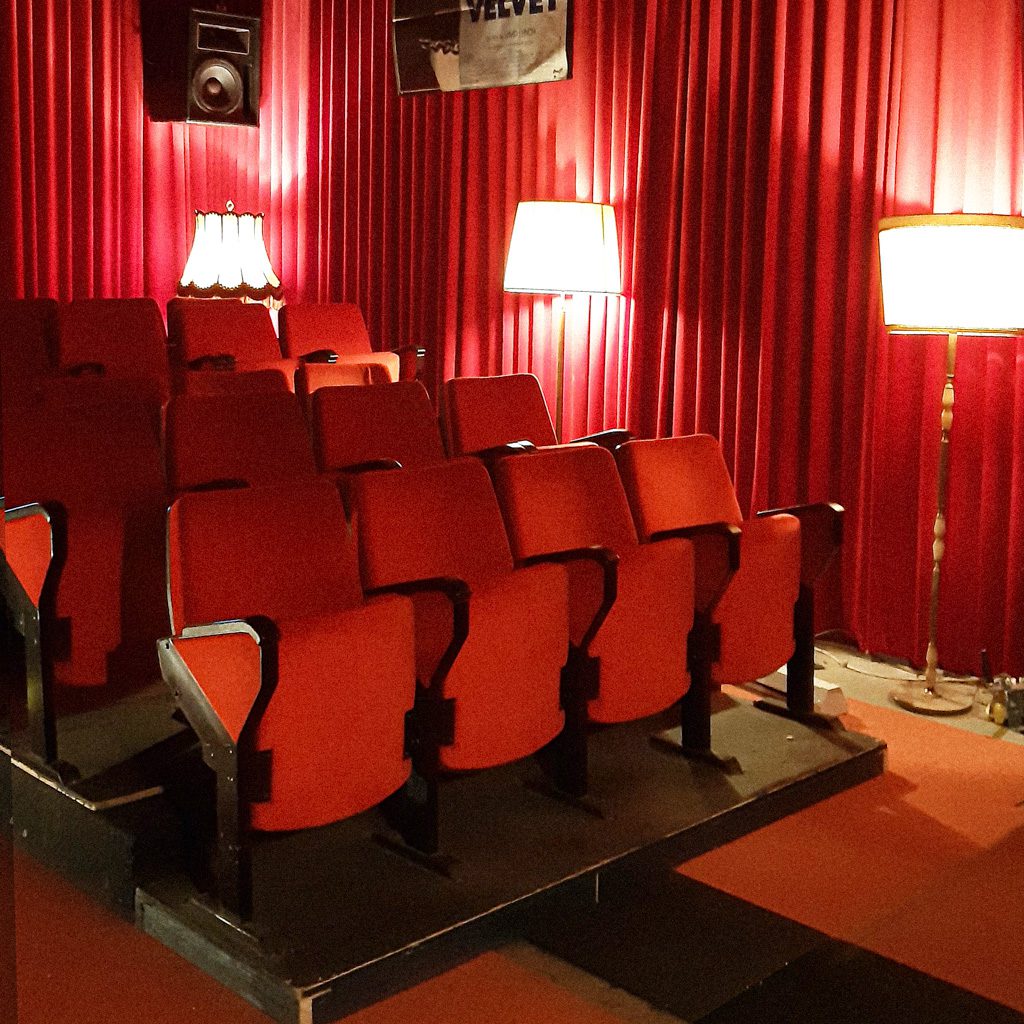 Picture of the cinema in Eva's studio. The picture is facing the seats, not the screen. The high walls are lined with long red curtains and the cinema seats are bright red as well.