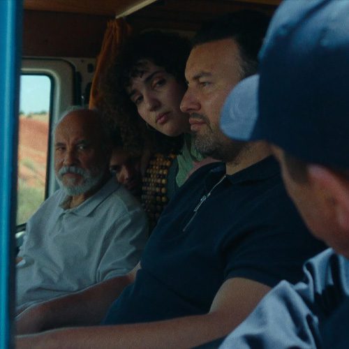 Square thumbnail for Porte Bagage. It's an over-shoulder medium shot, looking into the driver seat window of a blue van. In the van are three Dutch-Moroccan people. In the drivers seat is Farid, a middle aged man with a short beard. In the passenger seat is Musa, an old man with a white tunic on. Noor, a young woman with curly brown hair, is sticking her head in between the two seats and looking at the person looking into the window.