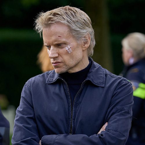 Square thumbnail from Van der Valk S4 E1 'Safe in Amsterdam'. It's a medium shot on Piet van der Valk. He's looking to the left side, brows furrowed and arms crossed. On his cheek is a cut with a bandage on it. Faintly visible in the background is a Dutch police officer.